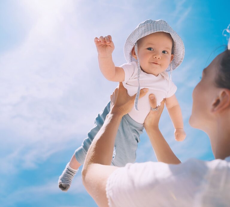 Junge Frau hebt lächelnd ein Baby mit gestreiftem Sonnenhut und weißem T-Shirt in den strahlend blauen Himmel. | © Getty Images/NataliaDeriabina