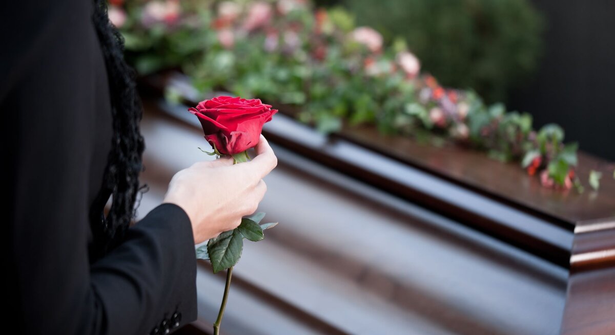 Eine Person in schwarzer Kleidung hält eine rote Rose vor einem mit Blumen geschmückten Sarg.