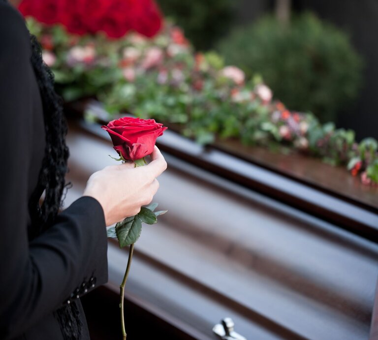 Eine Person in schwarzer Kleidung hält eine rote Rose vor einem mit Blumen geschmückten Sarg. | © Adobe Stock/Kzenon