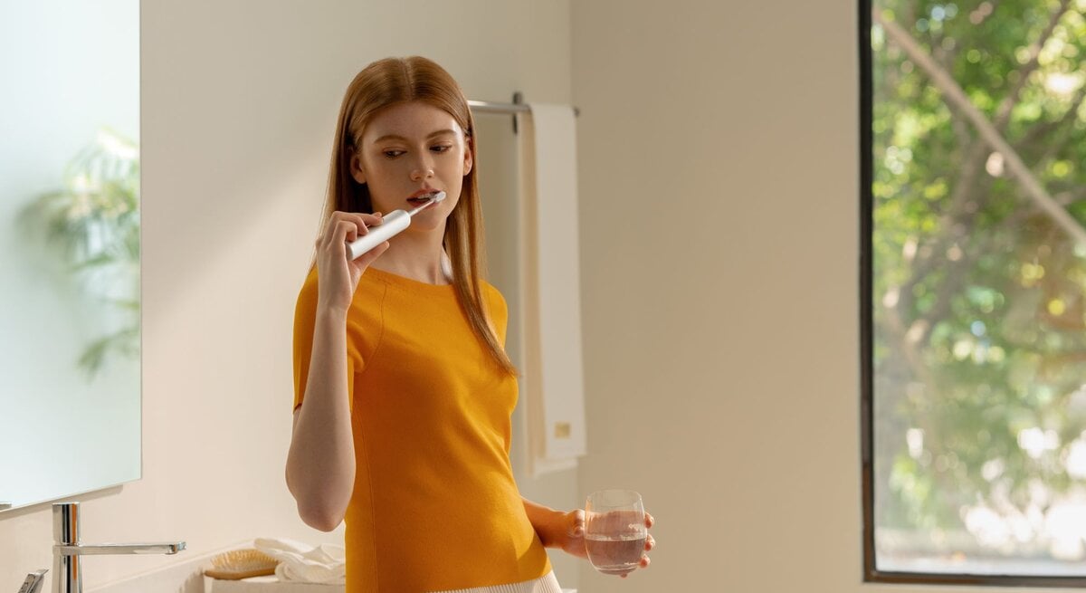 Junge Frau mit langen, roten Haaren putzt sich in einem modernen, hellen Badezimmer die Zähne, hält ein Glas Wasser.