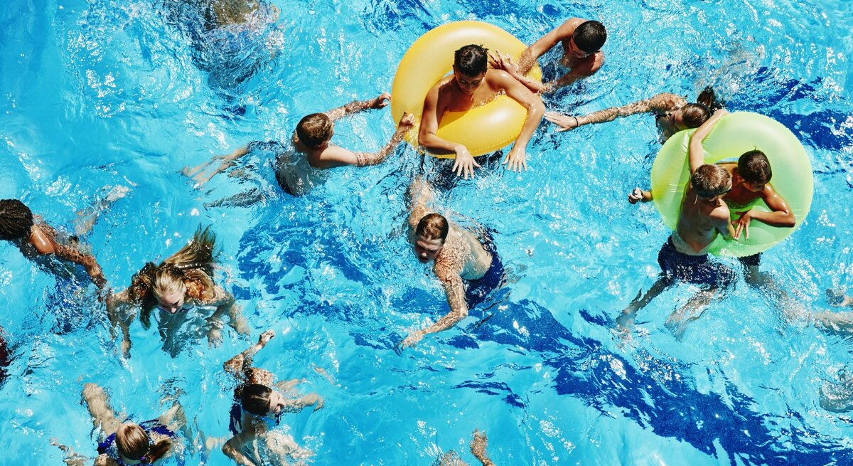 Mehrere Badegäste im Wasser eines Schwimmbeckens, auch Schwimmringe sind mit dabei, alle genießen sichtlich den sonnigen Tag.