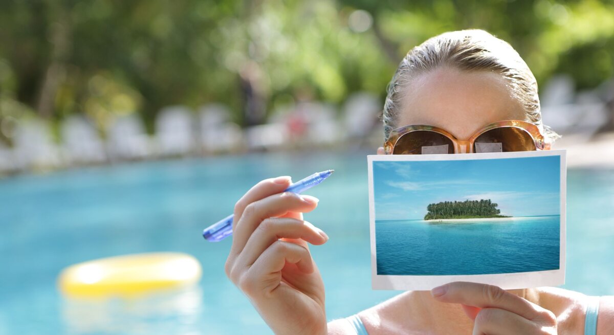 Eine Frau mit brauner Sonnenbrille hält eine Postkarte von einer Insel und einen blauen Stift vor einem Pool.