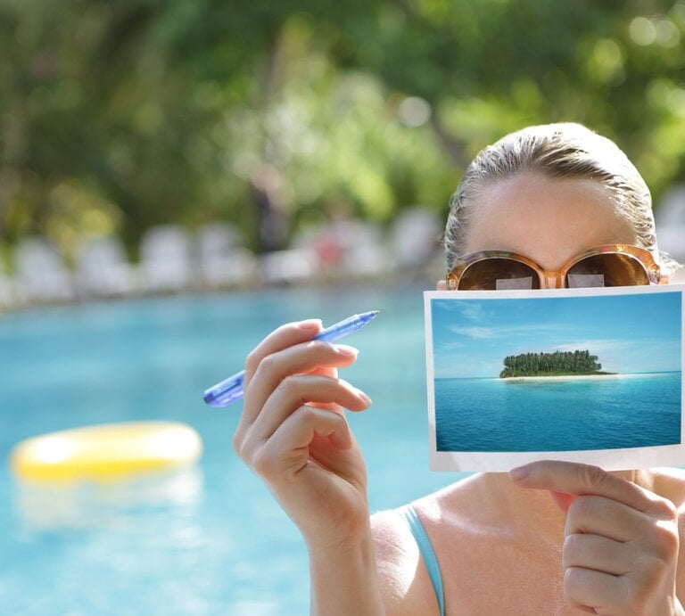 Eine Frau mit brauner Sonnenbrille hält eine Postkarte von einer Insel und einen blauen Stift vor einem Pool. | © Getty Images/Peter Cade