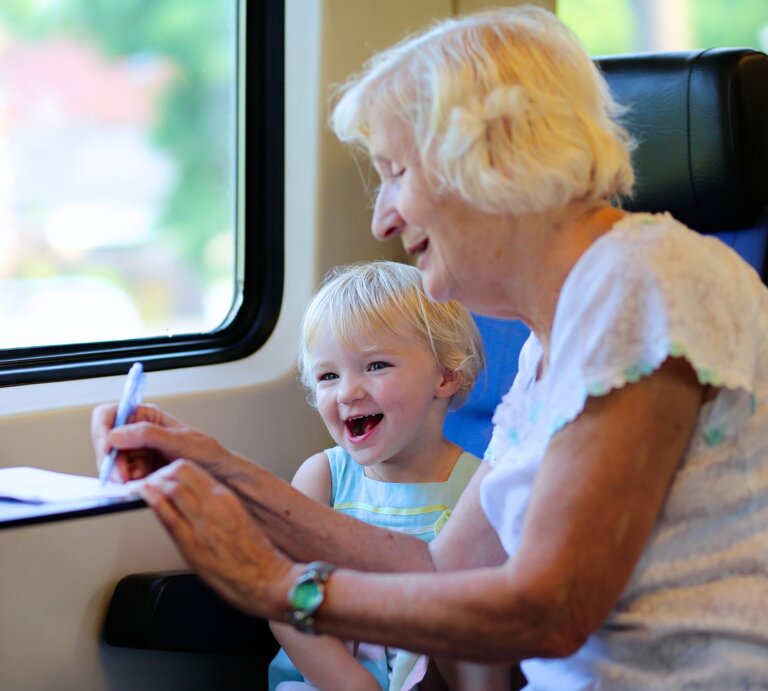 Lachendes blondes Kind sitzt neben einer fröhlichen Seniorin im Zug, die mit Stift und Papier auf dem Klapptisch schreibt. | © Shutterstock/ CroMary