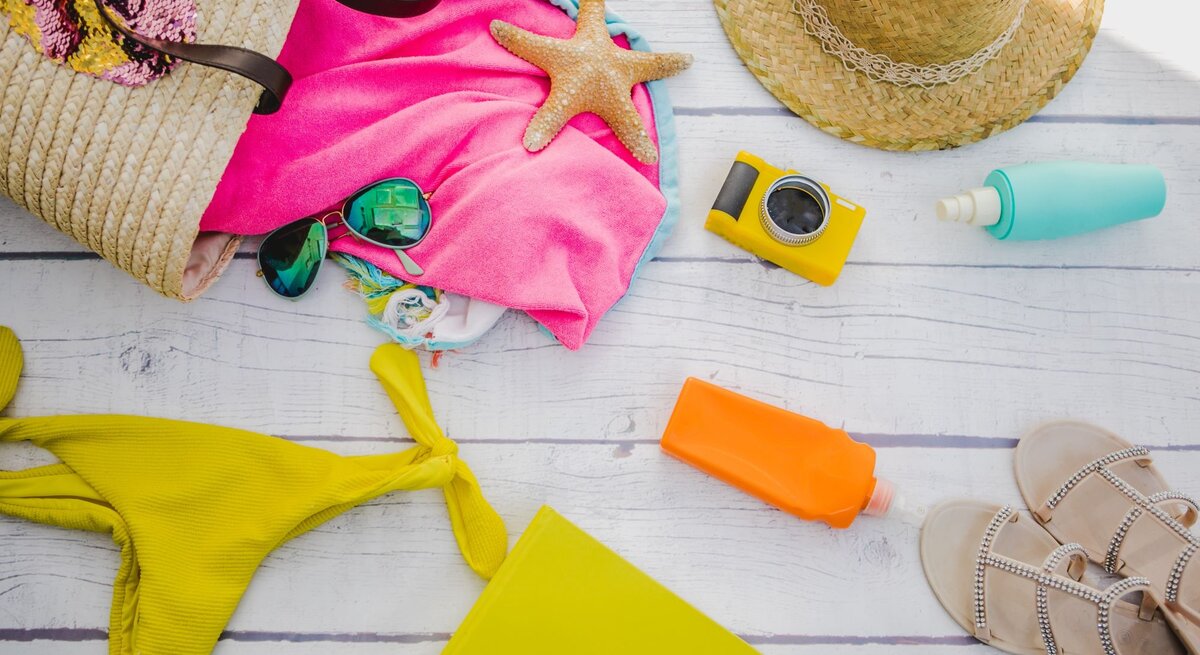 Eine gelbe Bikinihose, Sonnencreme, Kamera und Sonnenhut liegen auf einem Holztisch neben einer Strandtasche aus Stroh mit Handtuch und Sonnenbrille.