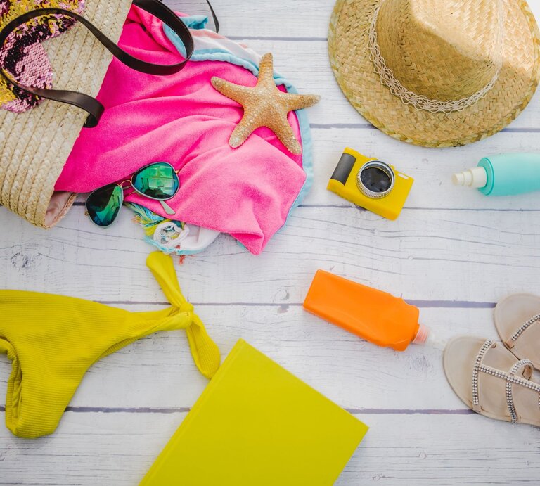 Eine gelbe Bikinihose, Sonnencreme, Kamera und Sonnenhut liegen auf einem Holztisch neben einer Strandtasche aus Stroh mit Handtuch und Sonnenbrille. | © Getty Images/Carol Yepes