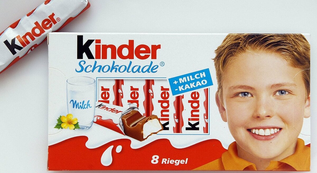 Packung Kinder Schokolade. Auf der linken Seite der Verpackung ist ein Glas Milch mit einem gelben Blümchen abgebildet, daneben sind einige Schokoladenriegel zu sehen. Ein einzelner Schokoladenriegel ist neben der Verpackung platziert. Auf der rechten Seite der Verpackung ist das lächelnde Gesicht eines Jungen zu sehen. 