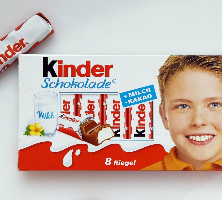 Packung Kinder Schokolade. Auf der linken Seite der Verpackung ist ein Glas Milch mit einem gelben Blümchen abgebildet, daneben sind einige Schokoladenriegel zu sehen. Ein einzelner Schokoladenriegel ist neben der Verpackung platziert. Auf der rechten Seite der Verpackung ist das lächelnde Gesicht eines Jungen zu sehen.  | © imago/Lars Reimann