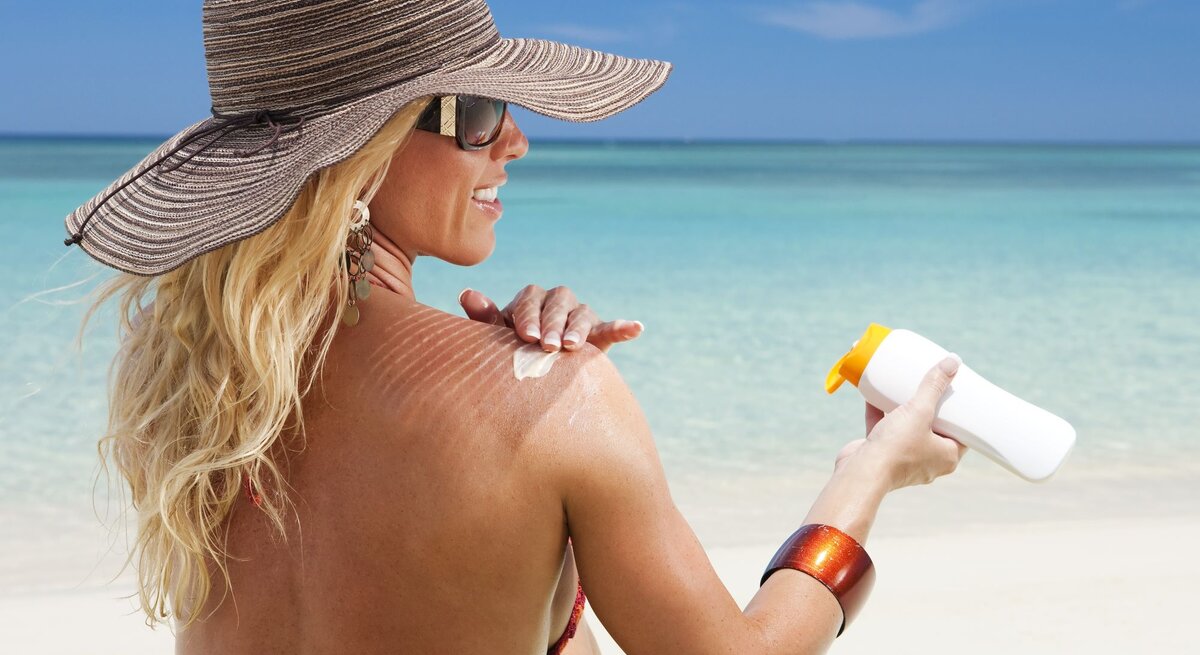 Eine Frau am Strand mit Hut und Bikini schmiert sich die Schulter mit Sonnencreme ein. Die Sonne scheint, der Himmel ist blau.