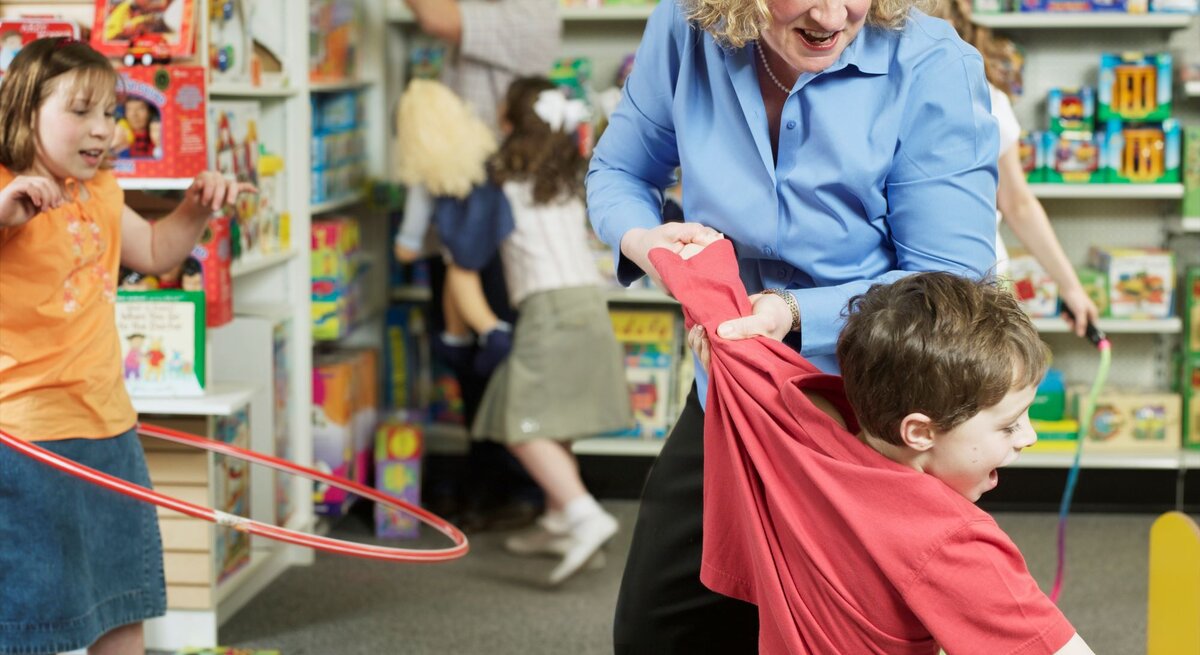 Ein Junge in rotem Shirt wird von einer blonden Frau in einem Spielzeugladen zurückgehalten, umgeben von spielenden Kindern und bunten Spielsachen.