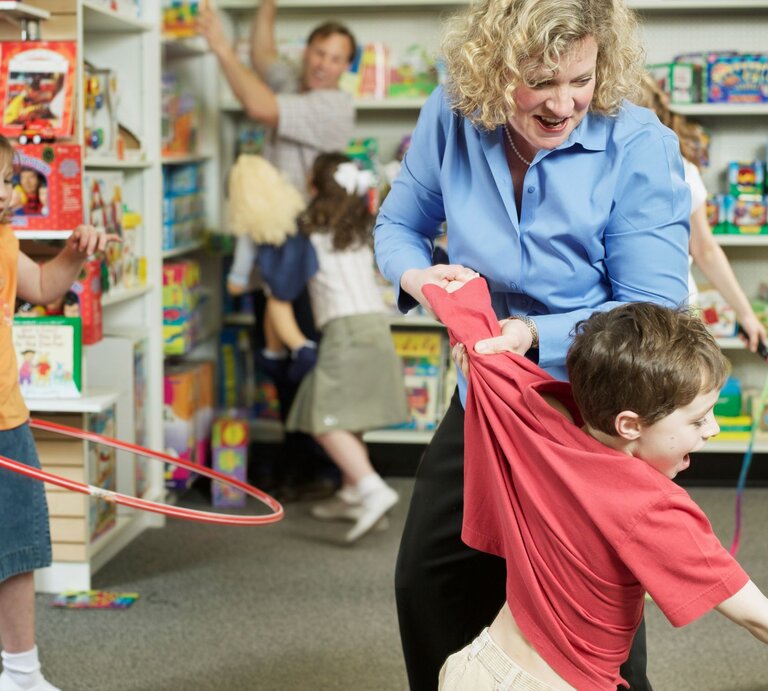 Ein Junge in rotem Shirt wird von einer blonden Frau in einem Spielzeugladen zurückgehalten, umgeben von spielenden Kindern und bunten Spielsachen. | © GettyImages/Sean Justice
