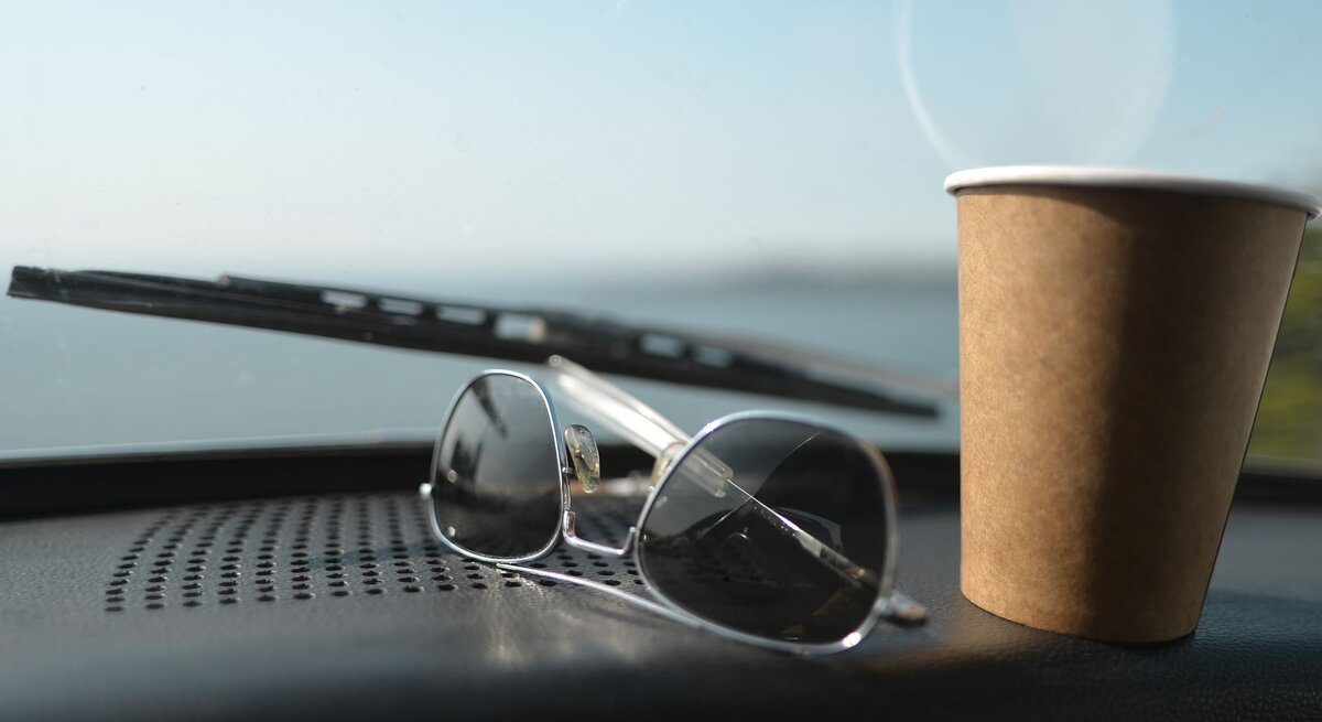 Sonnenbrille und brauner Pappbecher auf Armaturenbrett eines Autos mit Blick auf eine unscharfe Landschaft.