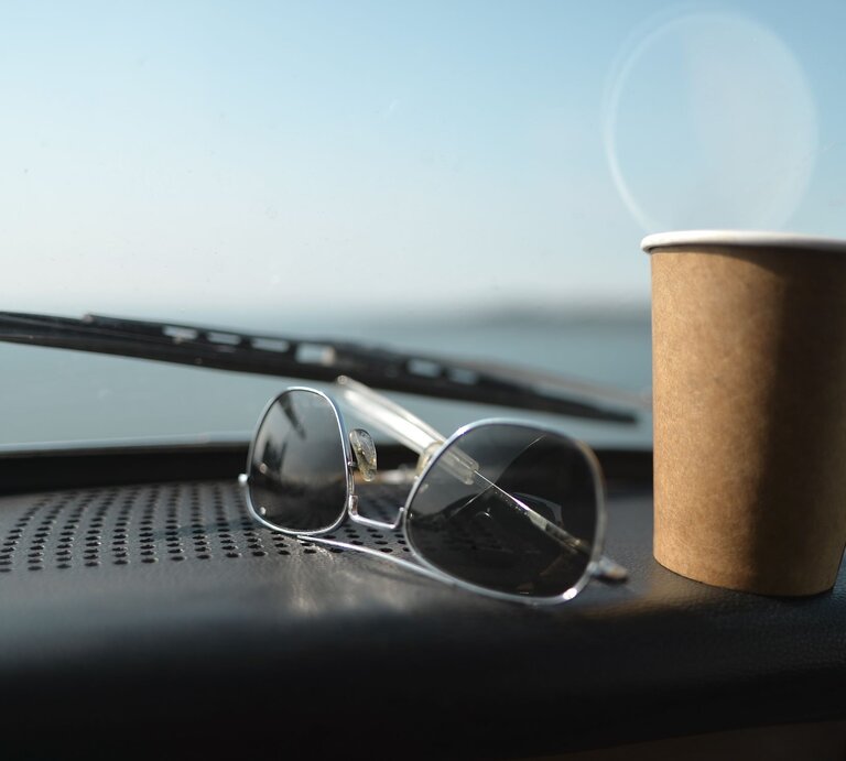 Sonnenbrille und brauner Pappbecher auf Armaturenbrett eines Autos mit Blick auf eine unscharfe Landschaft. | © GettyImages/robert reader