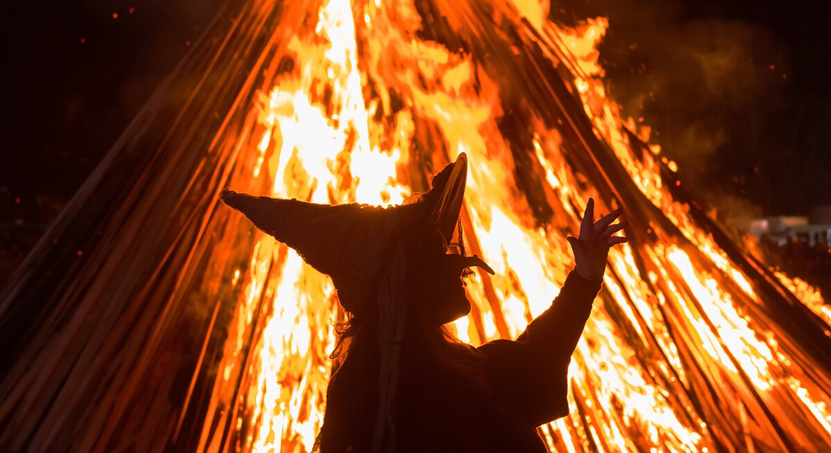 Zu sehen ist das Konterfei einer Hexe, die in der Nacht vor einem hellen, orangenen Feuer tanzt