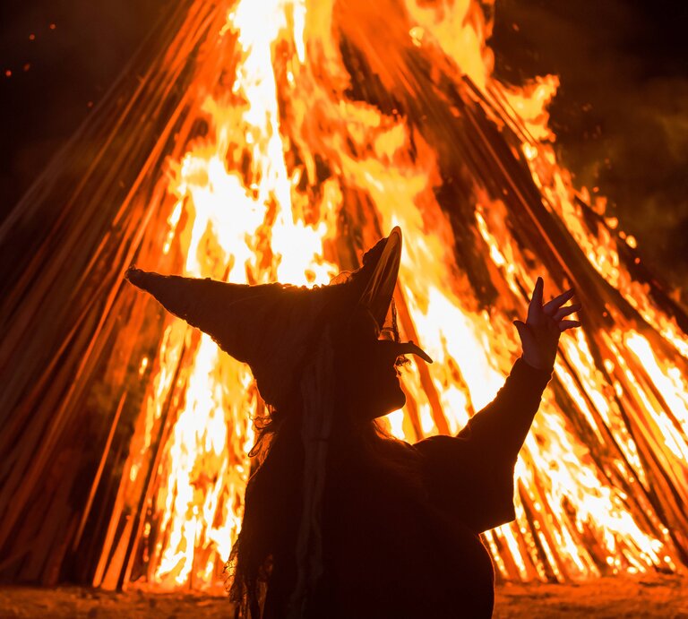 Zu sehen ist das Konterfei einer Hexe, die in der Nacht vor einem hellen, orangenen Feuer tanzt | © IMAGO / photo2000