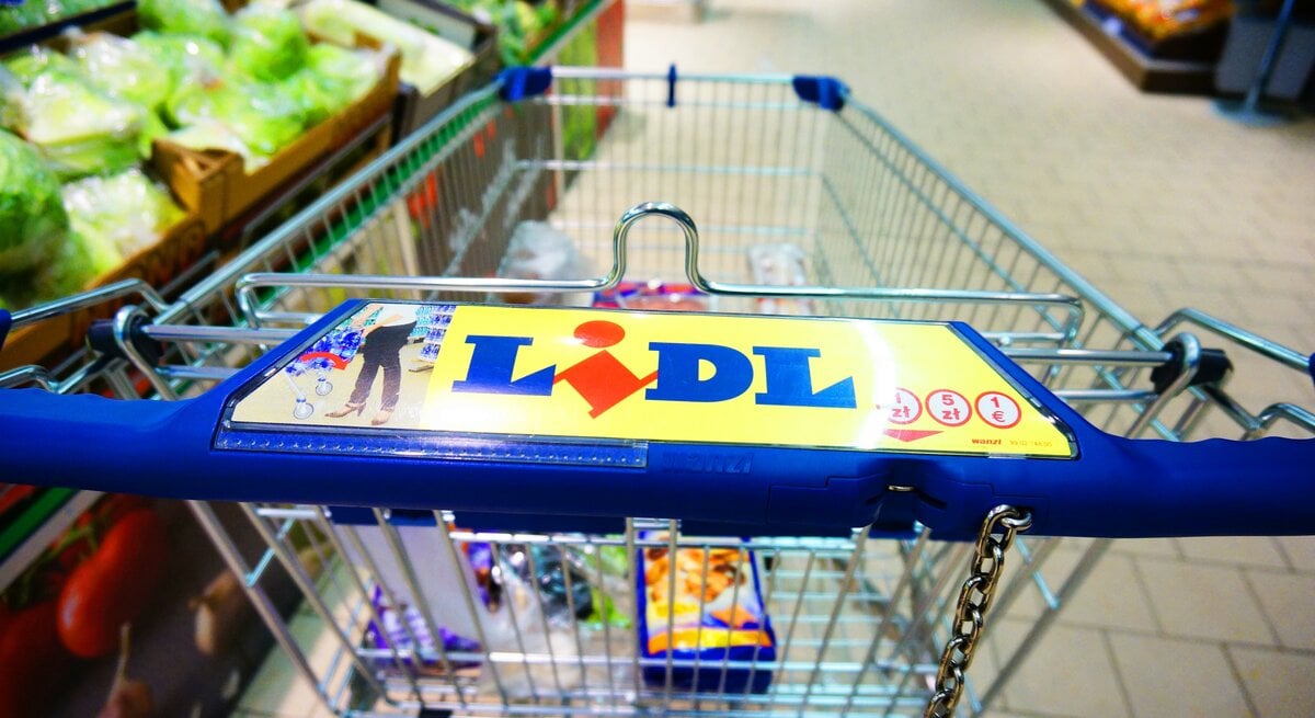 Einkaufswagen mit blauem Griff und Lidl-Logo im Supermarkt, im Hintergrund Gemüseabteilung und geflieste Böden sichtbar.