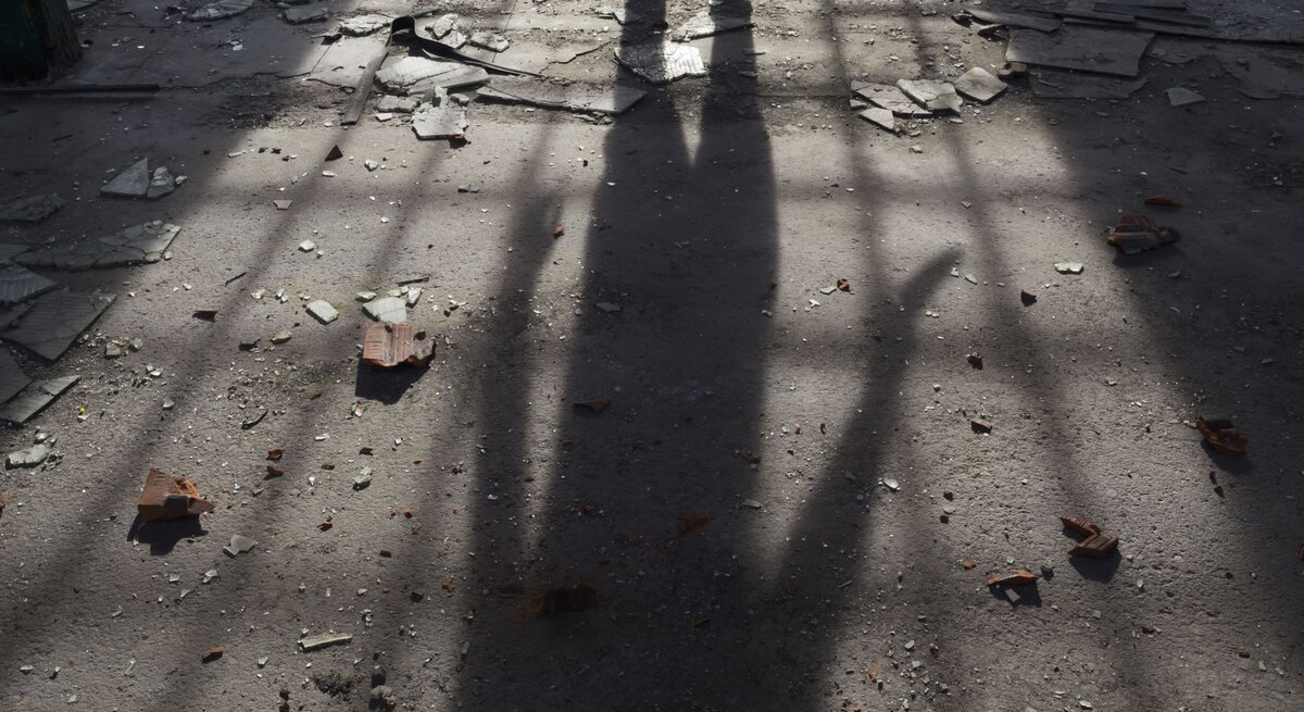  Der Schatten einer Person, die ein Messer hält, fällt bedrohlich auf einen mit Scherben übersäten Boden, was eine düstere und möglicherweise gefährliche Szene suggeriert, während das Tageslicht durch eine unbekannte Struktur ein düsteres Muster auf den Untergrund wirft.