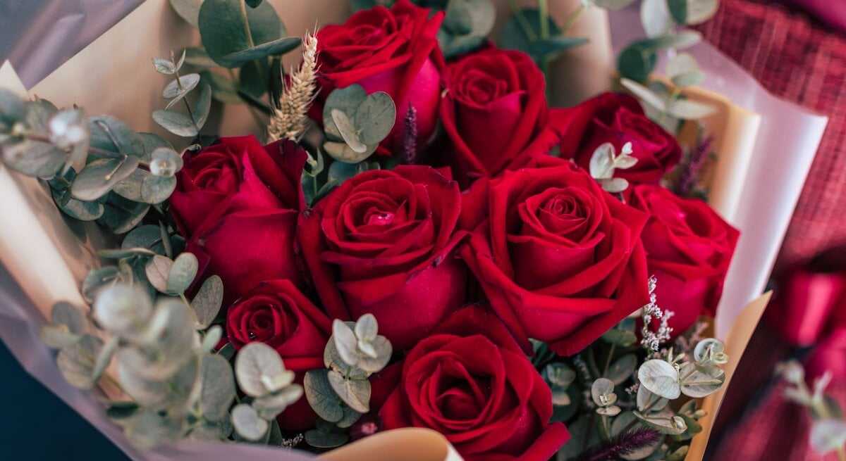 Ein üppiger Strauß aus leuchtend roten Rosen, umrahmt von Eukalyptuszweigen, in cremefarbenem Geschenkpapier kunstvoll arrangiert.
