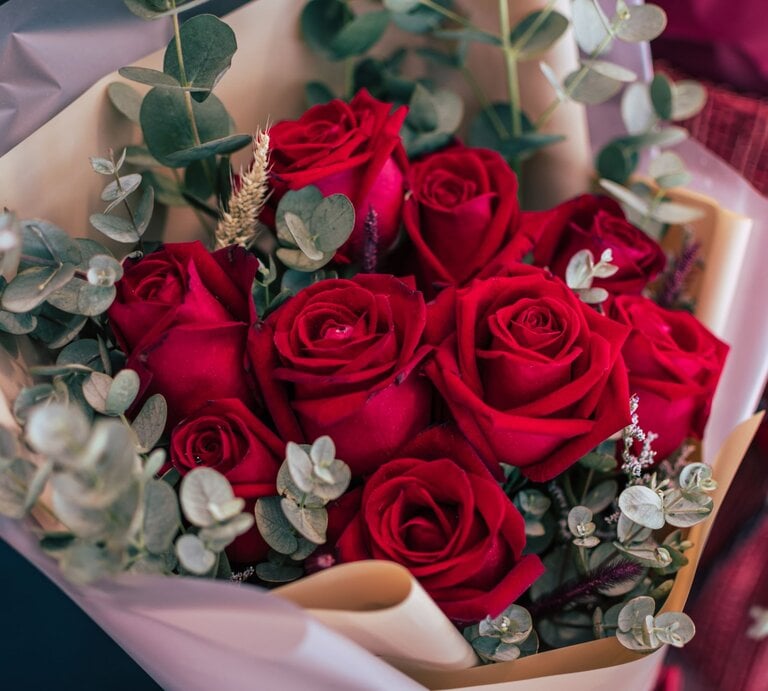 Ein üppiger Strauß aus leuchtend roten Rosen, umrahmt von Eukalyptuszweigen, in cremefarbenem Geschenkpapier kunstvoll arrangiert. | © GettyImages/sakchai vongsasiripat
