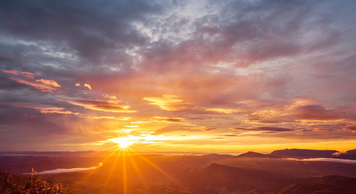 Ein atemberaubender Sonnenaufgang über einer hügeligen Landschaft, bei dem die Sonne gerade am Horizont erscheint. Der Himmel ist eine dramatische Leinwand aus orangen, gelben und blauen Farbtönen, durchzogen von den Strahlen der Sonne, die durch vereinzelte Wolken brechen und die Täler sowie den über dem Boden schwebenden Nebel erleuchten. Im Vordergrund zeigen sich Silhouetten von Pflanzen gegen den sich aufhellenden Himmel