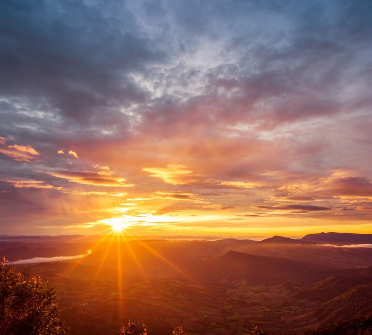 Ein atemberaubender Sonnenaufgang über einer hügeligen Landschaft, bei dem die Sonne gerade am Horizont erscheint. Der Himmel ist eine dramatische Leinwand aus orangen, gelben und blauen Farbtönen, durchzogen von den Strahlen der Sonne, die durch vereinzelte Wolken brechen und die Täler sowie den über dem Boden schwebenden Nebel erleuchten. Im Vordergrund zeigen sich Silhouetten von Pflanzen gegen den sich aufhellenden Himmel | © Adobe Stock/songdech17