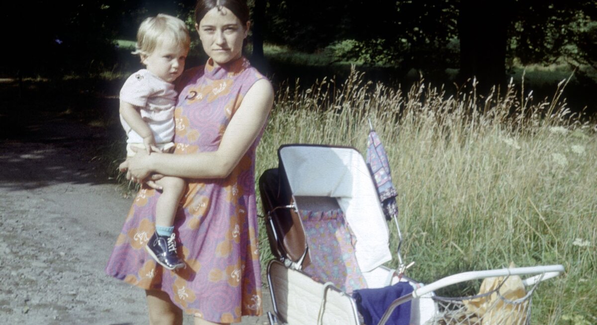 Mutter in lila Kleid mit Kind auf dem Arm, steht neben einem weißen Kinderwagen, im Hintergrund hohes Gras.