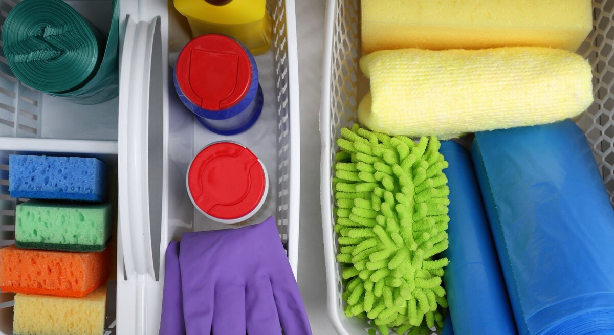 Geordnete Reinigungsmittel in weißen Körben: Schwämme, Müllbeutel, violette Handschuhe und Mikrofasertücher in Gelb und Grün.