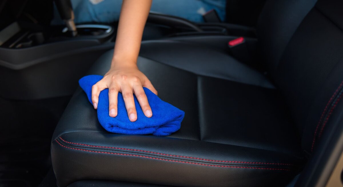 Person reinigt den Sitz eines Autos, verwendet dafür ein blaues Reinigungstuch und wischt über den schwarzen Ledersitz. Der Fokus liegt auf der Hand und dem Tuch, während der Rest des Autos nur teilweise sichtbar ist.