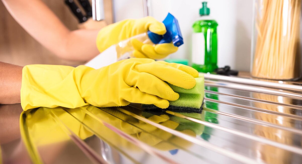 Zwei Hände in gelben Handschuhe putzen eine Edelstahlspüle mit einem Schwamm und Putzmittel.