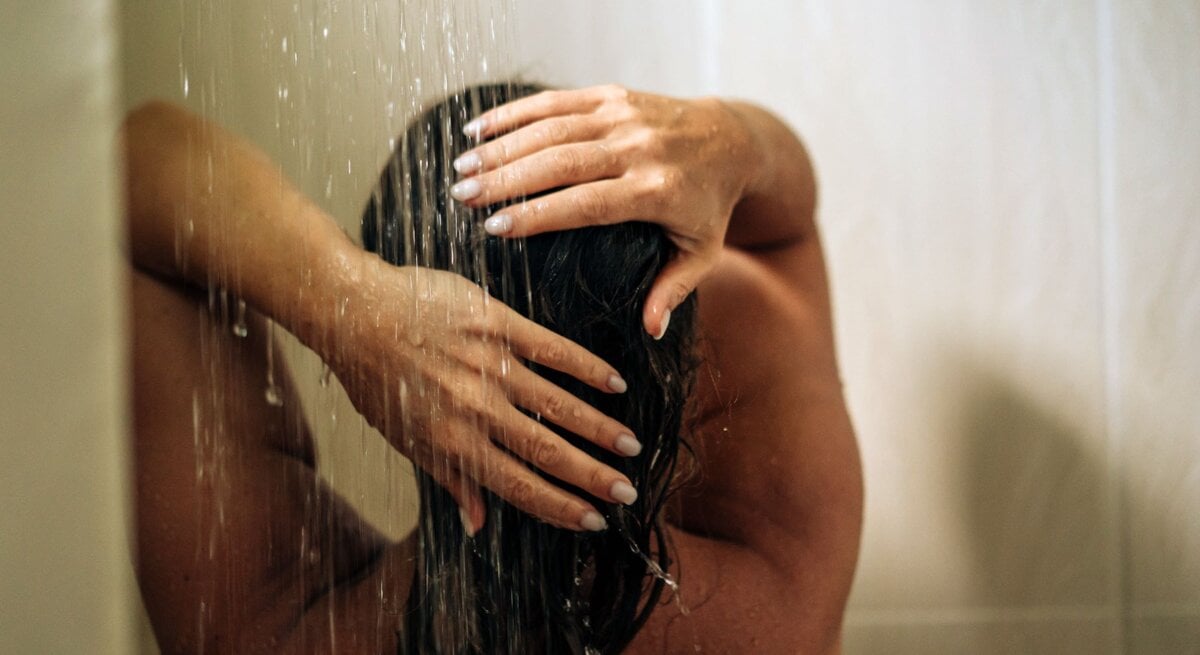 Eine Frau mit nassen Haaren steht in der Dusche und hält sich die Hände über den Kopf, während Wasser auf sie herabströmt.