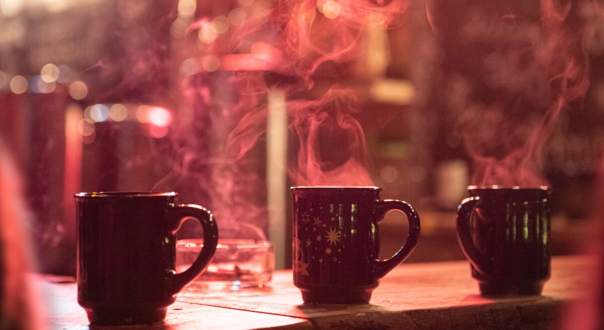 Drei dampfende Tassen Glühwein auf einem Holztisch, unscharfer Weihnachtsmarkt im Hintergrund mit Lichterglanz.