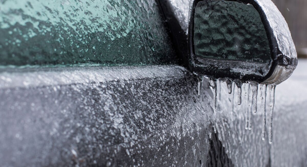 Außenspiegel und Fenster eines Autos komplett von einer dicken Eisschicht bedeckt, an der Eiszapfen hängen.
