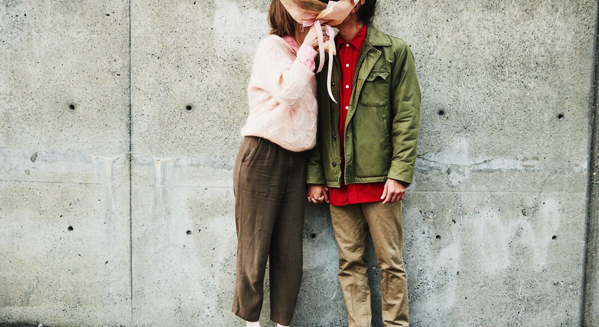 Das Bild zeigt ein Paar, das sich in einer liebevollen Pose gegenübersteht. Die Person auf der linken Seite trägt eine rosa Strickjacke, eine braune Hose und weiße Schuhe. Diese Person hält einen großen, bunten Blumenstrauß, der ihr Gesicht verdeckt. Die Person auf der rechten Seite trägt eine olivgrüne Jacke, ein rotes Hemd, braune Hosen und rote Turnschuhe. Auch das Gesicht dieser Person ist durch den Blumenstrauß verdeckt, was eine spielerische und romantische Atmosphäre schafft. Sie stehen vor einer Betonwand und auf einem hölzernen Untergrund, was einen interessanten Kontrast zur Wärme und Farbigkeit des Blumenstraußes bildet. Das Bild könnte symbolisch für die Anonymität und Universalität der Liebe stehen, oder es könnte einfach ein kreativer Ausdruck von Zuneigung sein.
