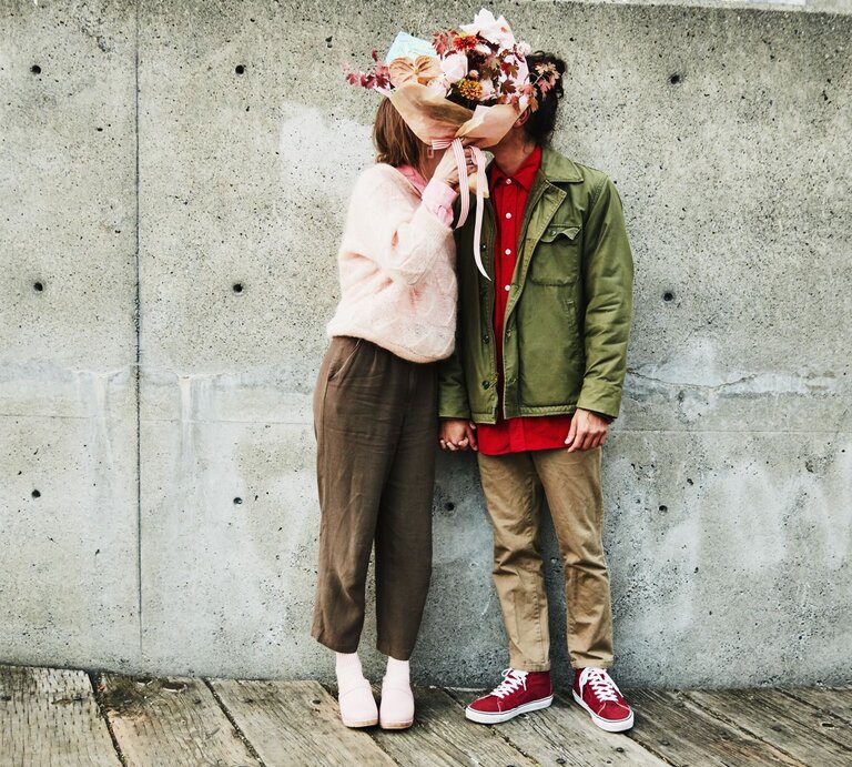 Das Bild zeigt ein Paar, das sich in einer liebevollen Pose gegenübersteht. Die Person auf der linken Seite trägt eine rosa Strickjacke, eine braune Hose und weiße Schuhe. Diese Person hält einen großen, bunten Blumenstrauß, der ihr Gesicht verdeckt. Die Person auf der rechten Seite trägt eine olivgrüne Jacke, ein rotes Hemd, braune Hosen und rote Turnschuhe. Auch das Gesicht dieser Person ist durch den Blumenstrauß verdeckt, was eine spielerische und romantische Atmosphäre schafft. Sie stehen vor einer Betonwand und auf einem hölzernen Untergrund, was einen interessanten Kontrast zur Wärme und Farbigkeit des Blumenstraußes bildet. Das Bild könnte symbolisch für die Anonymität und Universalität der Liebe stehen, oder es könnte einfach ein kreativer Ausdruck von Zuneigung sein. | © gettyimages/	Thomas Barwick