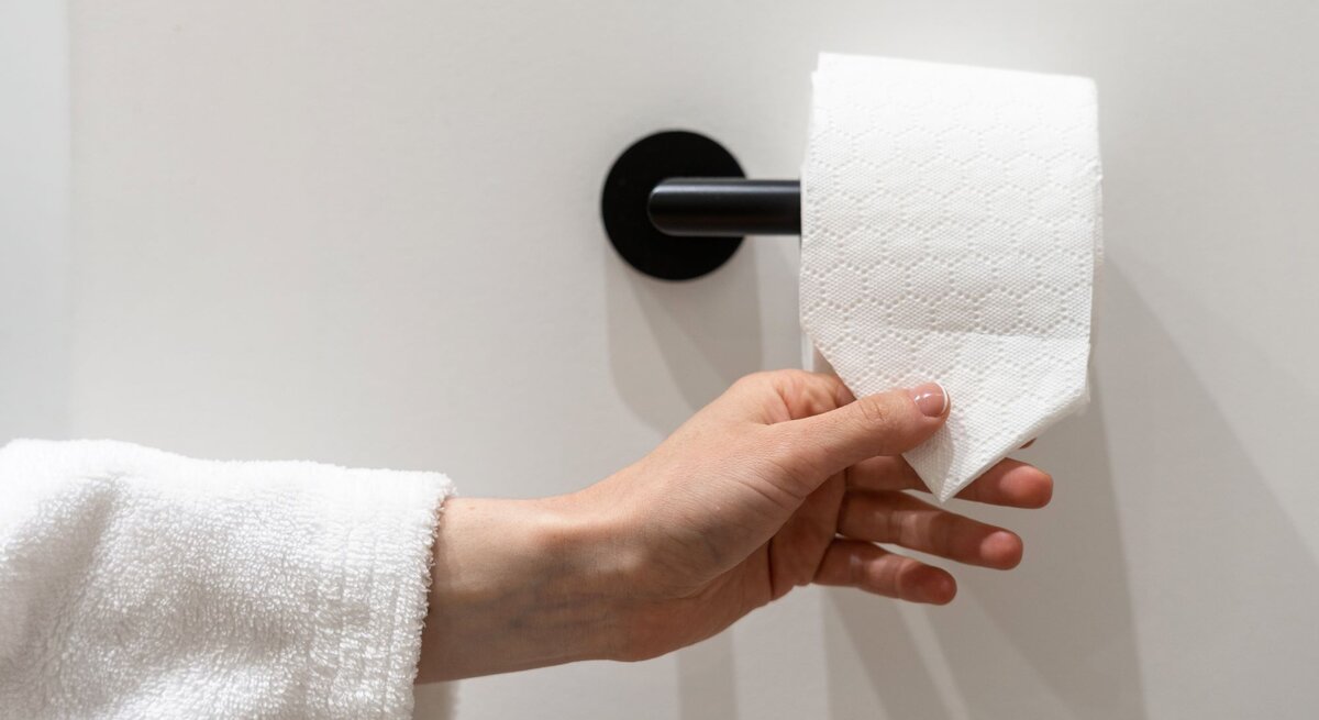 Eine Hand im weißen Bademantel greift nach einem dreieckig gefalteten Blatt Toilettenpapier an einer schwarzen Wandhalterung.