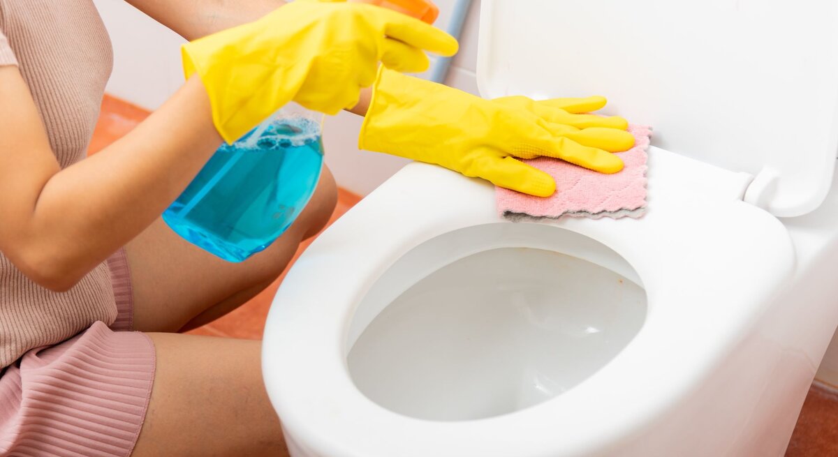 Zwei Hände in gelben Handschuhen reinigen eine Toilette und einen Toilettensitz.