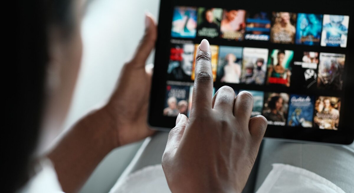 Eine Person mit dunkler Hautfarbe wählt auf einem Tablet im Querformat einen Film aus einer Streaming-Bibliothek aus.