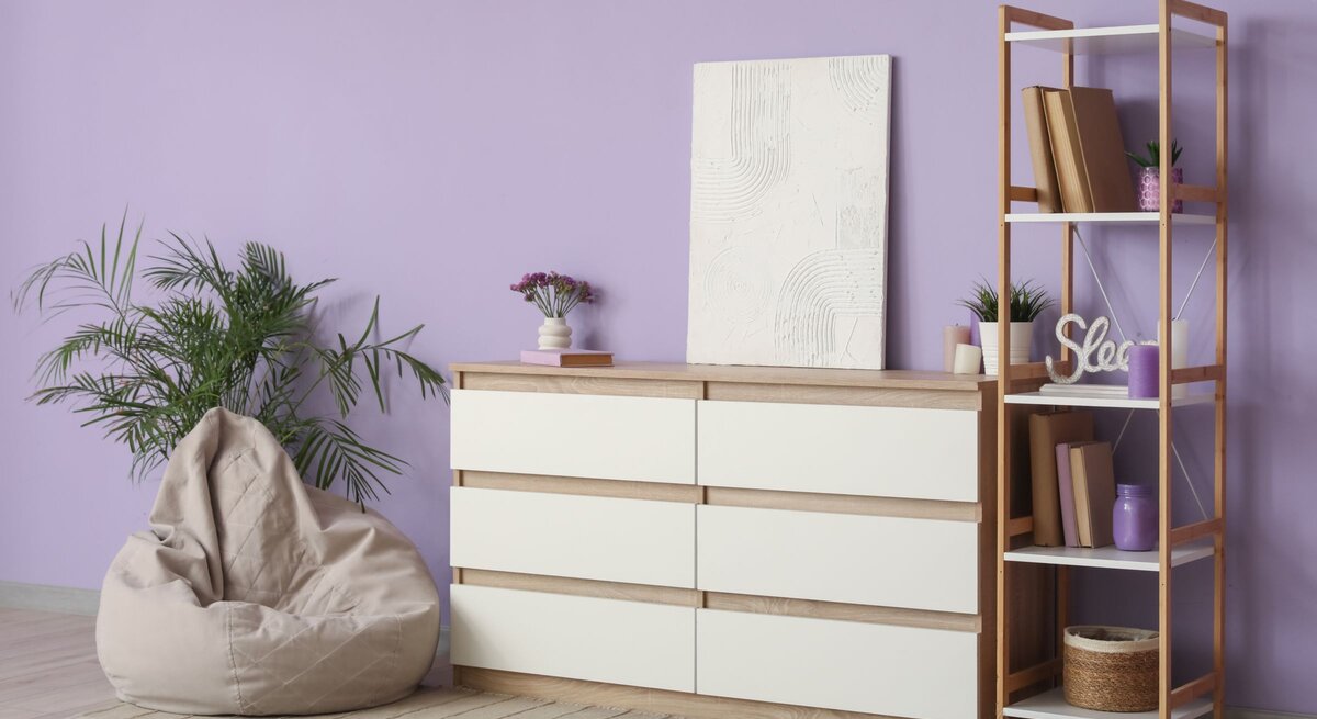 Modernes Wohnzimmer mit fliederfarbener Wand, Kommode aus Holz, Regal, Pflanzen, Sitzsack und ruhiger, heller Atmosphäre.