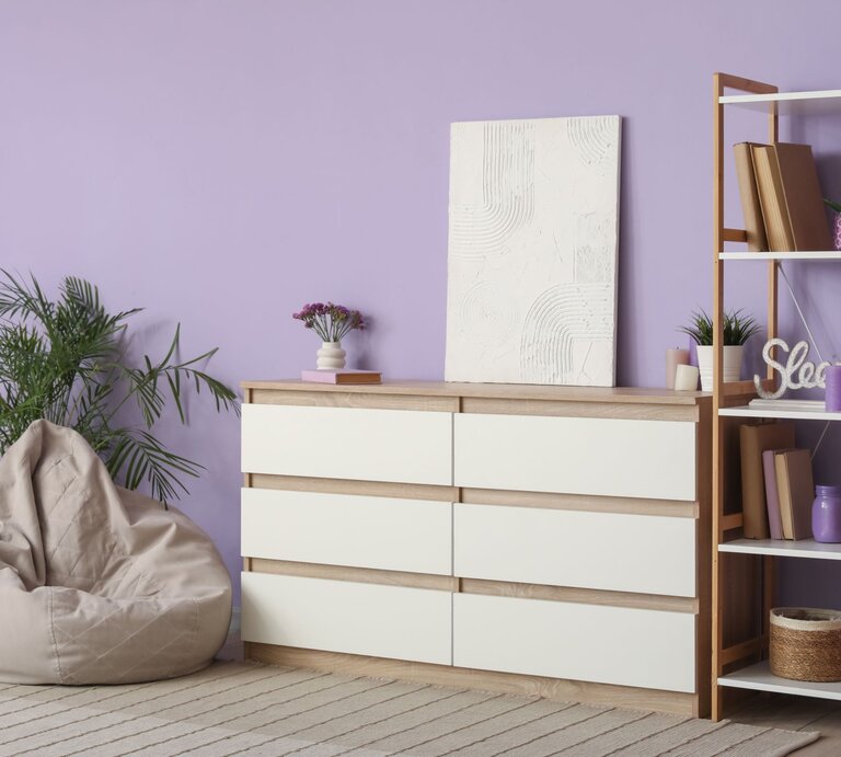 Modernes Wohnzimmer mit fliederfarbener Wand, Kommode aus Holz, Regal, Pflanzen, Sitzsack und ruhiger, heller Atmosphäre. | © Shutterstock/Pixel-Shot