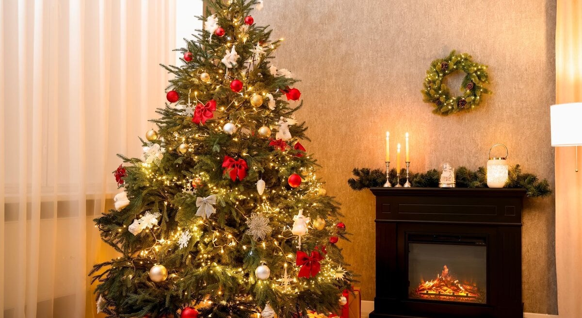 Geschmückter Weihnachtsbaum mit roten Schleifen und glänzenden Kugeln, daneben leuchtender Kamin mit Kerzen und Tannendekor.