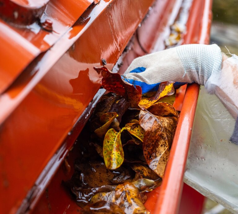 Nahaufnahme einer Hand mit Handschuh in einem roten Regenrinnensystem, entfernt herbstliche Blätter und Schmutz. | © Shutterstock/Natalia Kokhanova