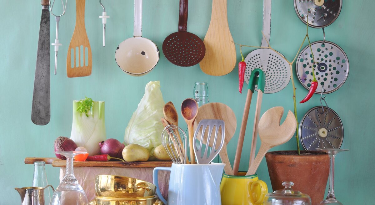 Küchenutensilien aus Holz, Glas und Metall sowie Gemüse wie Fenchel und Kohl arrangiert vor mintfarbenem Hintergrund auf einem Holztisch.