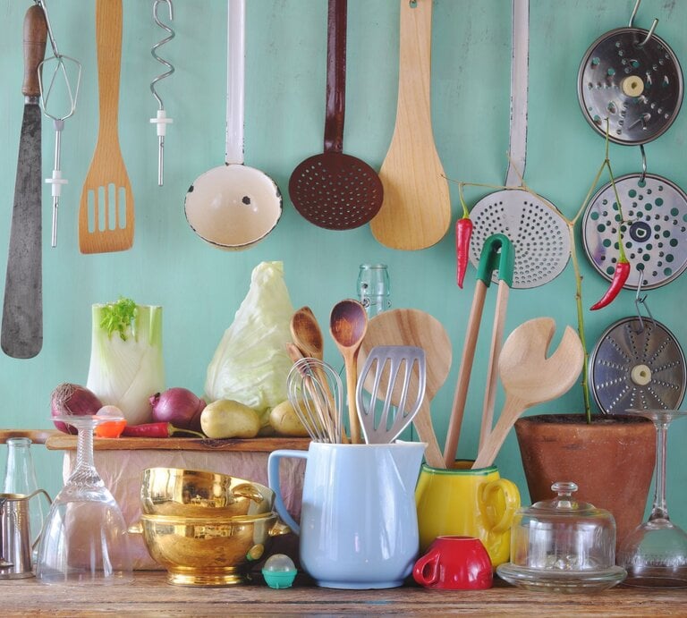 Küchenutensilien aus Holz, Glas und Metall sowie Gemüse wie Fenchel und Kohl arrangiert vor mintfarbenem Hintergrund auf einem Holztisch. | © Shutterstock/Thomas Bethge