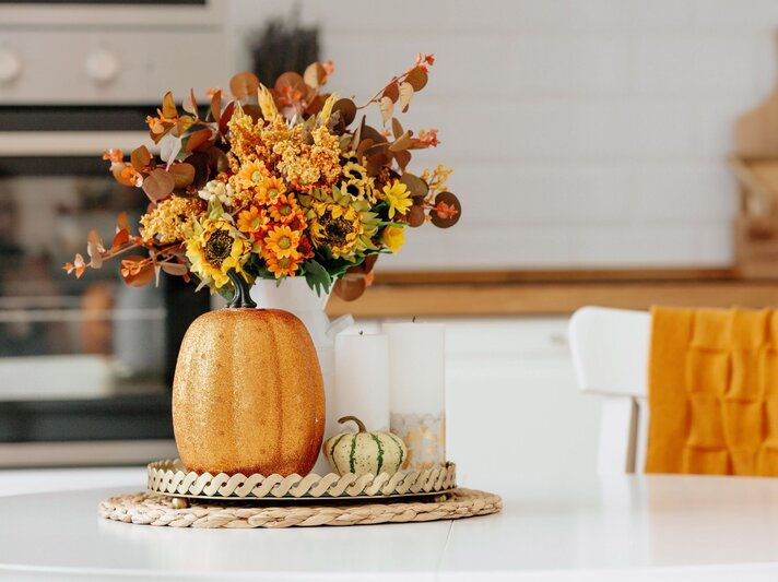 Ein goldener Deko-Kürbis, ein herbstlicher Blumenstrauß in warmen Farben und Kerzen stehen auf einem Tisch vor heller Küchenzeile. | © Shutterstock/PhotoJuli86