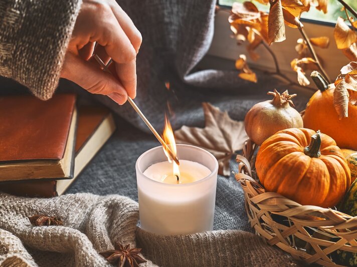 Eine Hand in Strickpullover entzündet mit einem Streichholz eine Kerze, daneben Kürbisse in einem Korb und Bücher auf grauem Hintergrund. | © Shutterstock/Caterina Trimarchi