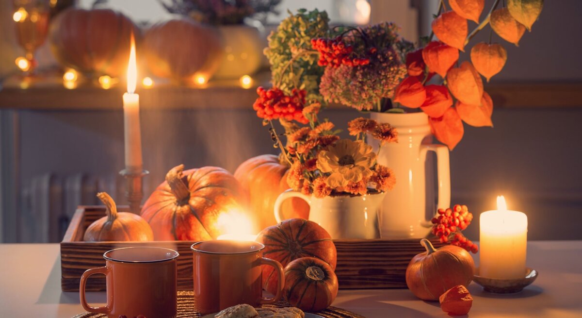 Stimmungsvolles Arrangement mit Kerzenlicht, orangefarbenen Kürbissen, herbstlichem Blumenstrauß und zwei Tassen Tee auf weißem Tisch.