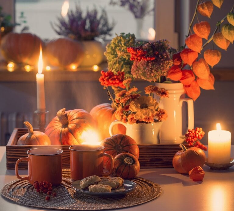 Stimmungsvolles Arrangement mit Kerzenlicht, orangefarbenen Kürbissen, herbstlichem Blumenstrauß und zwei Tassen Tee auf weißem Tisch. | © Shutterstock/Maya Kruchankova