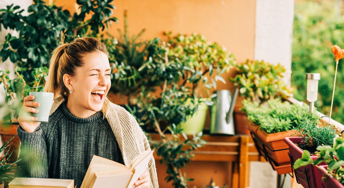 Junge blonde Frau in Strickpullover lacht mit Buch und Tasse auf grün bepflanztem Balkon voller Kräuter und Blumentöpfe.