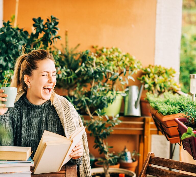 Junge blonde Frau in Strickpullover lacht mit Buch und Tasse auf grün bepflanztem Balkon voller Kräuter und Blumentöpfe. | © Adobe Stock / annanahabed