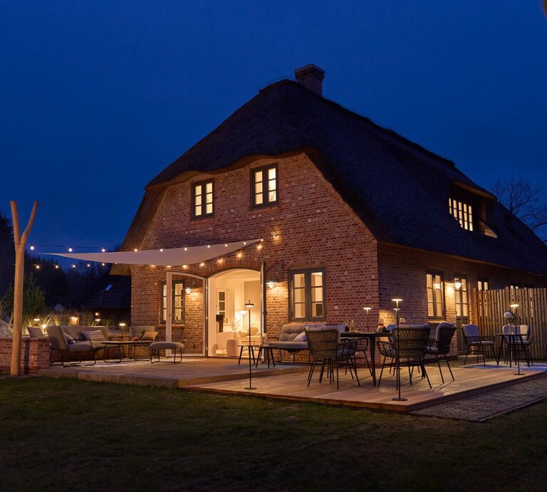 Gemütliches Reetdachhaus aus rotem Backstein bei Nacht, mit stimmungsvoll beleuchteter Holzterrasse und stilvollen Gartenmöbeln. | © LOTTO24